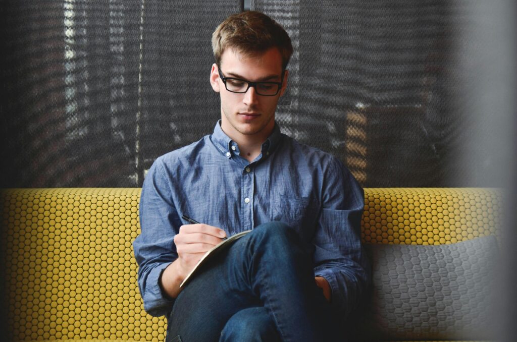 Home A young man in glasses writes in a notebook while sitting on a stylish couch indoors.
